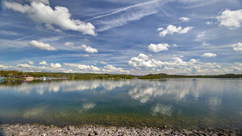 Carsington water harbour reflection polarised This landscape photograph taken in the late morning during spring shows Carsington Water, a reservoir located in the Peak District. The image captures the lake with clear, polarised reflections of clouds and sky on its surface, creating a striking mirror effect. Along the shoreline, the water's clarity reveals the stones beneath, and several boats are visible moored near the harbour, contributing to the scene's tranquil atmosphere. The surrounding landscape includes green hills and wooded areas, while the sky overhead is marked by fluffy white clouds and contrails. Carsington Water is featured prominently as the main subject, with its expanse and reflective qualities providing a distinctive visual characteristic typical of reservoirs in this region.